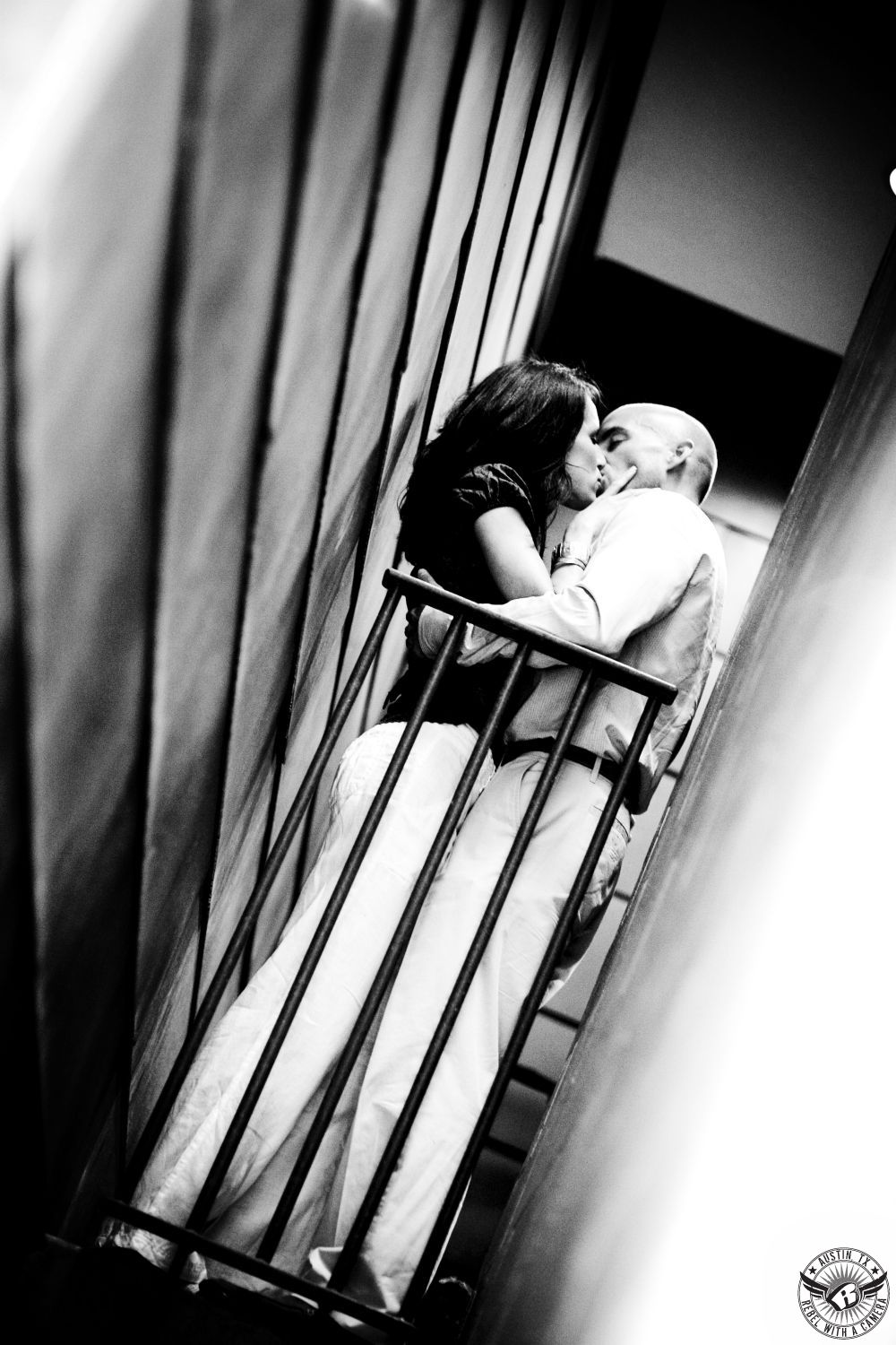 Hot, curvy, brunette wearing a black tight tank top and flowing white pants passionately kisses a guy wearing a grey dress shirt  and white pants standing against a black rail in a thin ally way with metal siding on the walls near SOCO in this daring black and ahite engagement image shot from below in Austin.  